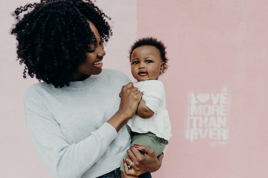 young and happy black mom stands in front of a pink wall with her baby girl.