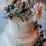 Two tier wedding cake topped with greenery and a few large pastel pink flowers running down the side of the textured cake.