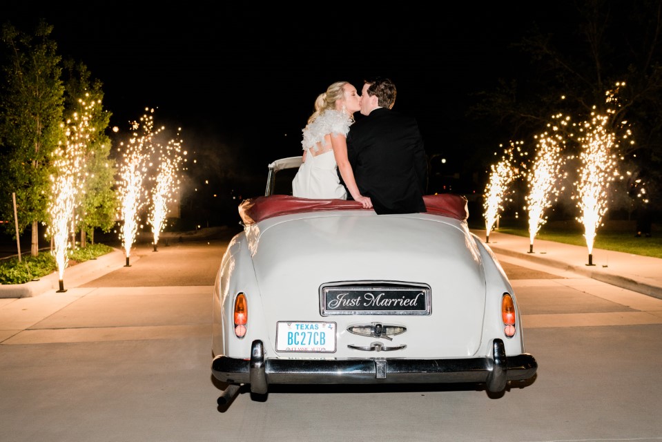 Newlyweds drive off in a classic car as fireworks ignite on both sides of the road