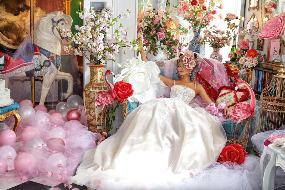 Model wears sleeveless Ines DiSanto Spring 2023 wedding dress ballgown while sprawled across a blue velvet chair surrounded by whimsical and colorful details such as flamingo statues and pink balloons.